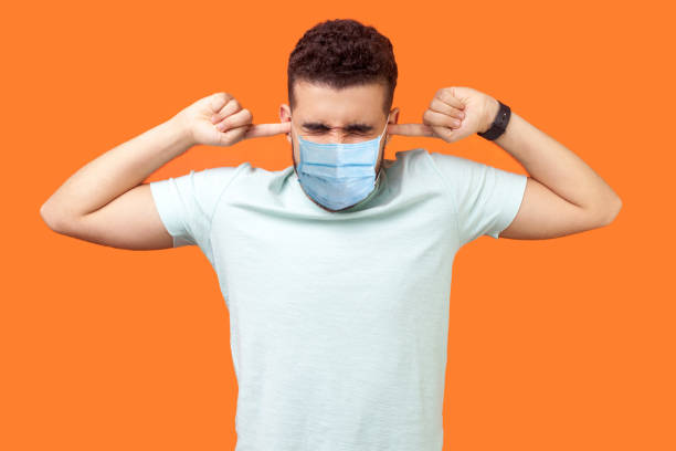 Don't want to listen anymore. Portrait of man standing with medical mask irritated grimace, tightly covering his ears not to hear annoying loud sound. Don't want to listen anymore. Portrait of man standing with medical mask irritated grimace, tightly covering his ears not to hear annoying loud sound. indoor studio shot isolated on orange background man closing ears photos stock pictures, royalty-free photos & images