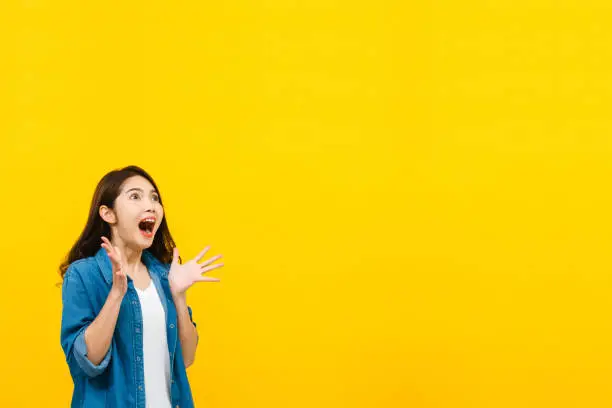 Photo of Shocked young Asian woman with hands on cheeks gasping and looking up to copy space in hand studio shot isolated on yellow background.