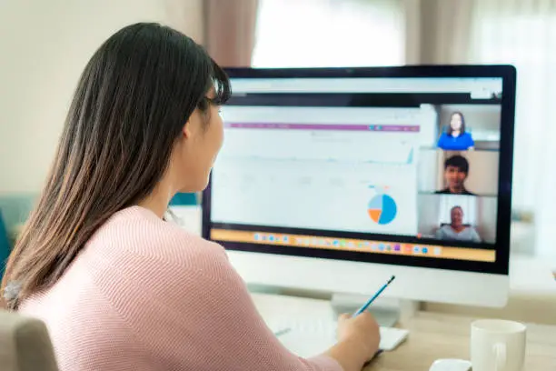 Photo of Back view of Asian business woman talking to her colleagues about plan in video conference. Multiethnic business team using computer for a online meeting in video call. Group of people smart working from home.