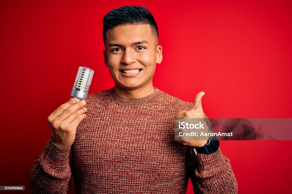 Young handsome latin man holding led lightbulb over isolated red background happy with big smile doing ok sign, thumb up with fingers, excellent sign Cut Out Stock Photo