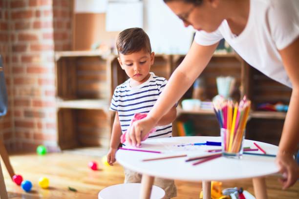 istock Beautiful teacher and toddler boy drawing draw using colored pencils at kindergarten 1214093329