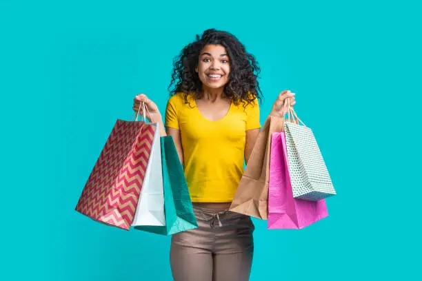 Cute and funny dark skinned girl holding piles of shopping bags on both hands isolated over blue background. Shopping, sales, black friday concept.