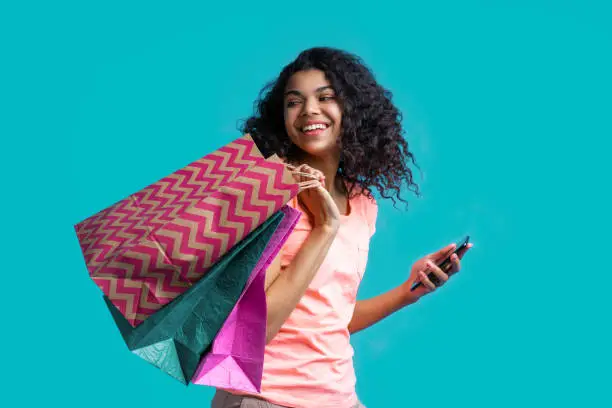 Cute african american girl with perfect smile posing with a pile of paper shopping bags and mobile phone in hands. Online shopping concept.