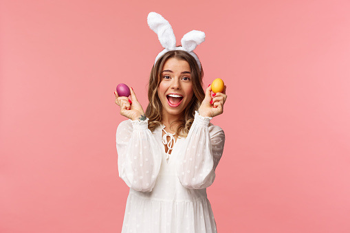 Holidays, spring and party concept. Portrait of excited charming young woman celebrating Easter in rabbit ears and white party dress, dancing with two painted eggs, pink background.