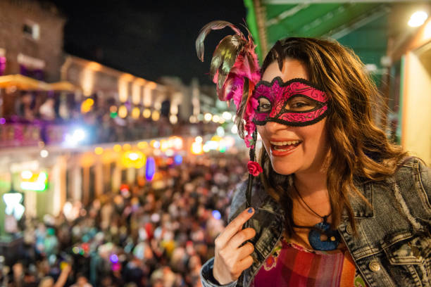 This is a photograph at night of a masked Caucasian woman in her 30’s enjoying the scene on Bourbon Street in New Orleans from a balcony view during Mardi Gras.