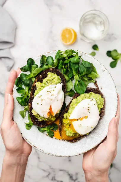 Avocado and poached egg on rye bread. Female hands holding healthy vegetarian breakfast or snack rye toast with mashed avocado, greends and egg