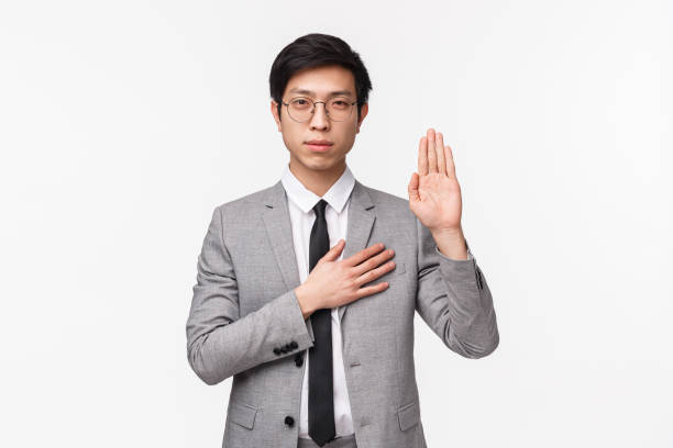 retrato de cintura arriba de joven asiático devoto y guapo en traje gris, haciendo promesa, promesa o juramento, levante una mano y ponga la palma de la mano en oído como honesto y sincero, fondo blanco - waistup fotografías e imágenes de stock