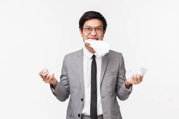 Waist-up portrait of annoyed, pissed-off mad young asian male office worker, eating paper, grimacing aggressive and distressed, ripping paper, tearing up report, standing white background.