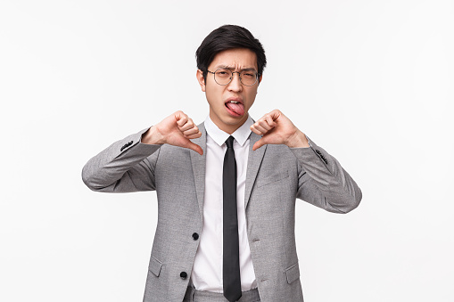 Waist-up portrait of skeptical, disappointed and unsatisfied asian businessman in grey suit, showing tongue and thumbs-down, squinting shaking head in rejection, no it is bad, white background.