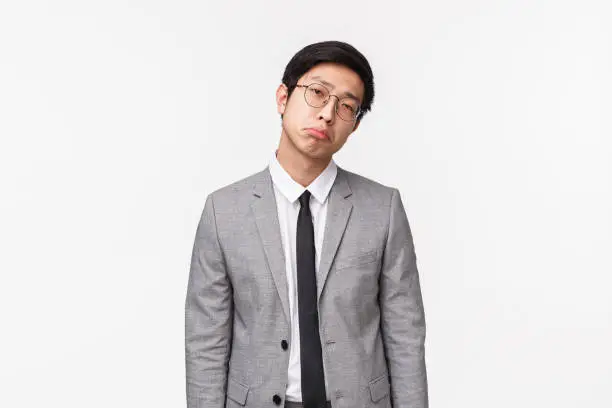 Waist-up portrait of gloomy and reluctant, unimpressed bored asian man in suit, businessman pulling unsatisfied grimace, looking judgemental and skeptical camera, standing white background.