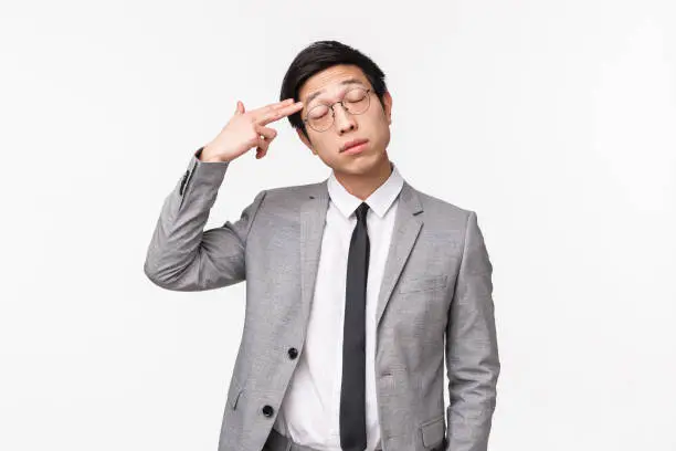 Waist-up portrait of annoyed and fed-up, tired asian guy in grey suit, close eyes and making gun gesture over forehead, as if shooting himself reacting to stupid annoying talk, white background.