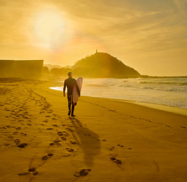 Zurriola Beach at sunset. San Sebastian, Basque Country. Spain. A surfer walking on the Zurriola Beach at sunset with the Monte Urgull in the background. San Sebastian, Basque Country, Guipuzcoa. Spain. neoprene photos stock pictures, royalty-free photos & images