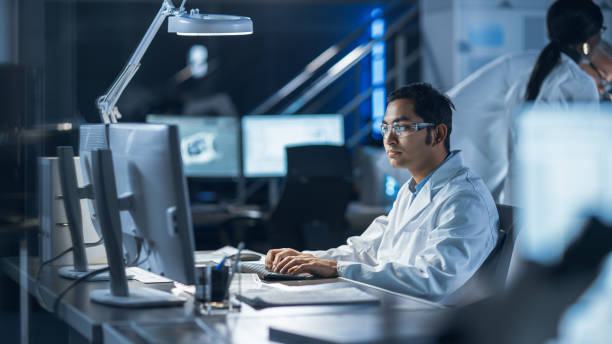 istock Male IT Scientist Uses Computer Showing System Monitoring and Controlling Program. In the Background Technology Development Laboratory with Scientists, Engineers Working 1214111307