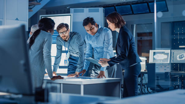 istock Engineers Meeting in Technology Research Laboratory: Engineers, Scientists and Developers Gathered Around Illuminated Conference Table, Talking and Finding Solution. Industrial Design Facility 1214111362