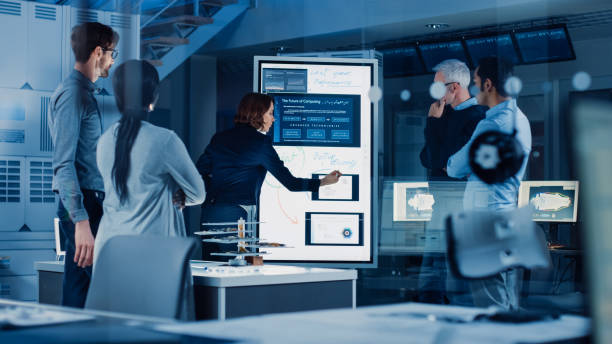 istock Engineers Meeting in Technology Research Laboratory: Female Engineer Leads Presentation Writes on Interactive Digital Whiteboard, Shows Machine Blueprint, Data Analytics and Neural Network while Colleagues Listening 1214111383