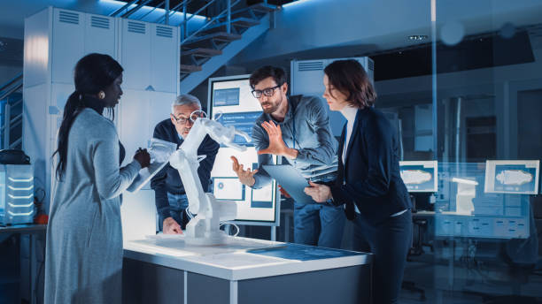 istock Engineers Meeting in Robotic Research Laboratory: Engineers, Scientists and Developers Gathered Around Illuminated Conference Table, Talking, Using Tablet and Analysing Design of Industrial Robot Arm 1214111404