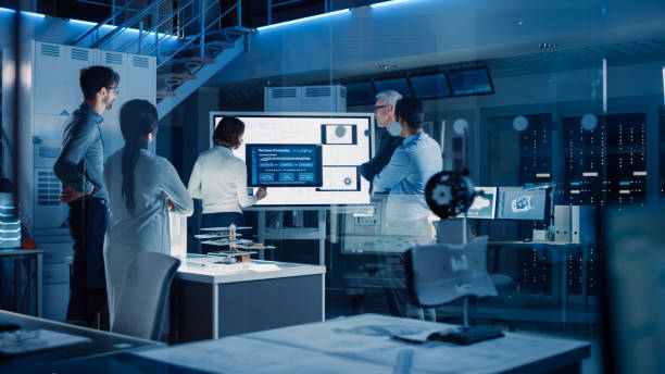 istock Engineers Meeting in Technology Research Laboratory: Female Engineer Leads Presentation Using Vertical Digital Whiteboard, Shows Machine Blueprint, Data Analytics and Neural Network while Colleagues Listening 1214111409