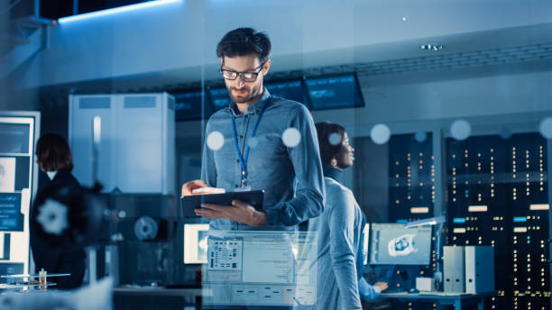 In Technology Research Facility: Chief Engineer Stands in the Middle of the Lab and Uses Tablet Computer. Team of Industrial Engineers, Developers Work on Engine Design Use Digital Whiteboard and Computers In Technology Research Facility: Chief Engineer Stands in the Middle of the Lab and Uses Tablet Computer. Team of Industrial Engineers, Developers Work on Engine Design Use Digital Whiteboard and Computers computer equipment stock pictures, royalty-free photos & images