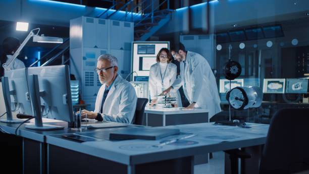 istock Diverse International Team of Industrial Scientists and Engineers Wearing White Coats Working on Heavy Machinery Design in Research Laboratory. Professionals Using Computers and Talking 1214111421