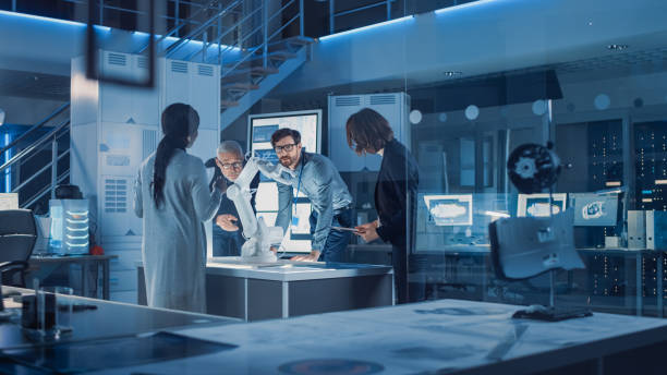 istock Engineers Meeting in Robotic Research Laboratory: Engineers, Scientists and Developers Gathered Around Illuminated Conference Table, Talking, Using Tablet and Analysing Design of Industrial Robot Arm 1214111423