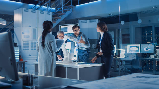 istock Engineers Meeting in Robotic Research Laboratory: Engineers, Scientists and Developers Gathered Around Illuminated Conference Table, Talking, Using Tablet and Analysing Design of Industrial Robot Arm 1214111450