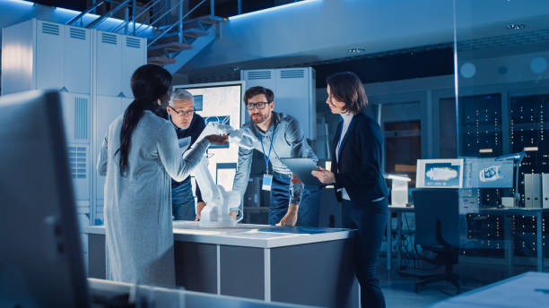 istock Engineers Meeting in Robotic Research Laboratory: Engineers, Scientists and Developers Gathered Around Illuminated Conference Table, Talking, Using Tablet and Analysing Design of Industrial Robot Arm 1214111467
