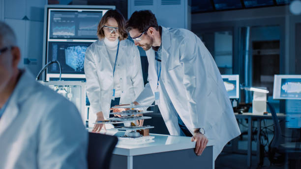 istock Shot in Industrial Design Research Laboratory, Specialist Uses Microscope, Designer Works on Personal Computer, Female Scientists Writes on Glass Whiteboard, Team of Developers Work with 3D Printer and Motherboard. 1214112099