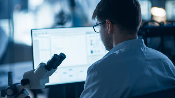 istock Shot of a Male IT Scientist Uses Computer with Electronic Microscope. In the Background Technology Development Laboratory with Scientists, Engineers Working 1214112153
