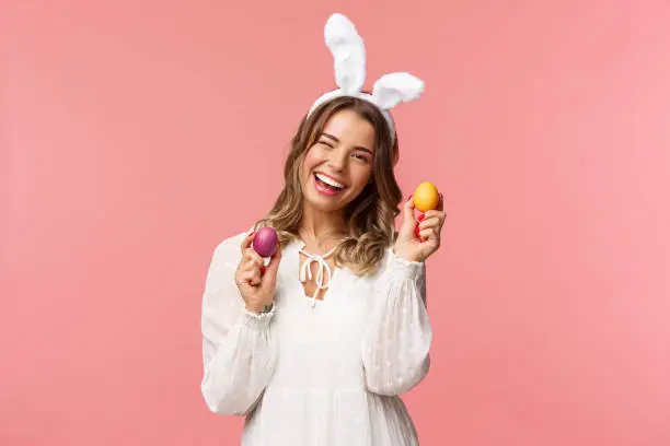 Photo of Holidays, spring and party concept. Cheerful good-looking blond woman celebrating Easter day in rabbit ears, holding two painted eggs and wink camera, smiling happily, pink background