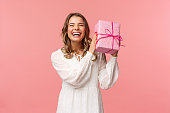 Holidays, celebration and women concept. Portrait of happy charismatic blond girl shaking gift box wondering whats inside as celebrating birthday, receive b-day presents, pink background