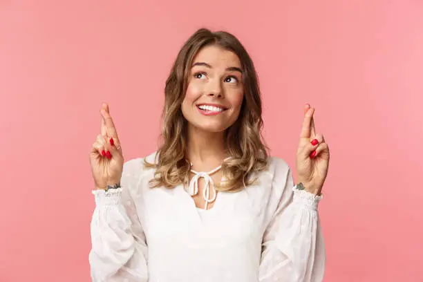 Photo of Close-up portrait of hopeful, optimistic attractive woman in white dress, bite lip and smiling as daydreaming, hope dream come true, making wish or praying, standing pink background