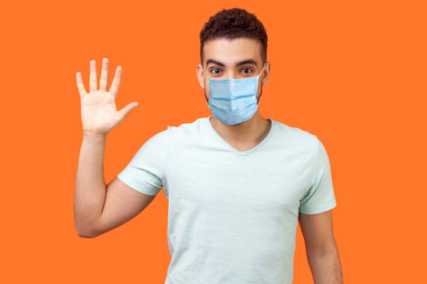 Hello! Portrait of friendly glad handsome man with medical mask in casual white t-shirt showing hi gesture with waving hand and smiling sincerely. Hello! Portrait of friendly glad handsome man with medical mask in casual white t-shirt showing hi gesture with waving hand and smiling sincerely. indoor studio shot isolated on orange background i 5 stock pictures, royalty-free photos & images