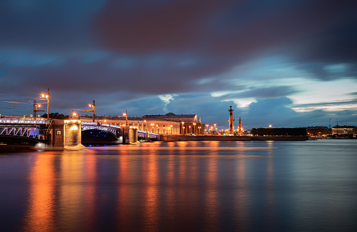 Beautiful white night view on illuminated Palace Bridge, Neva embankment and raster columns. All of them are famous landmarks of St Petersburg city, Russia. Long exposure photo. Dramatic cloudscape.