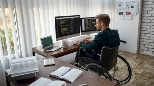 istock Full concentration. Back view of young professional web developer in a wheelchair writing program code on multiple computer screens while sitting at his workplace in the modern office 1214133669