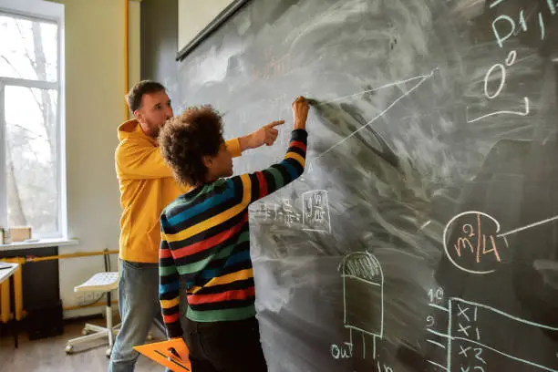 Photo of The great aim of education is not knowledge but action. Young male science teacher explaining main rules and formulas in physics and mechanics. A boy is drawing on the blackboard