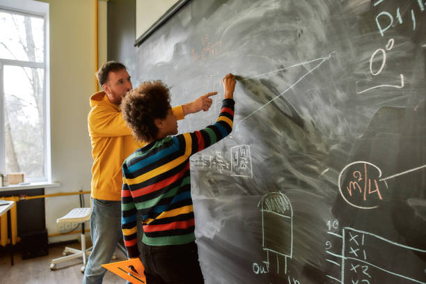 o grande objetivo da educação não é o conhecimento, mas a ação. jovem professor de ciências explicando as principais regras e fórmulas em física e mecânica. um garoto está desenhando no quadro negro - mathematics mathematical symbol blackboard education - fotografias e filmes do acervo