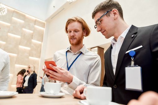 Businesspeople interacting, looking at the screen of smartphone while having coffee break at business conference. Business, communication concept. Horizontal shot. Selective focus