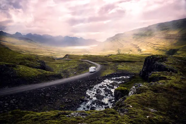 white camper van driving along winding mountain road through beautiful sunlit valley surrounded by mountains, Iceland