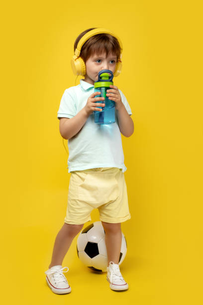 photo du petit garçon sérieux retenant le ballon de football et écoutant la musique dans le casque au-dessus du fond jaune. - water child bottle little boys photos et images de collection