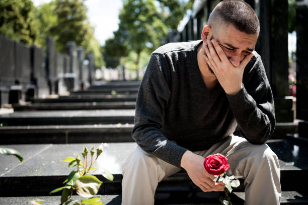 грустный и подавленный вдовец в черной одежде, стоящий на коленях перед надгробием, держащий цветок и стонующий из-за потери семьи. концепц� - moaning стоковые фото и изображения