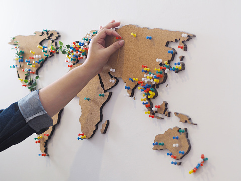 Woman hand putting red pin and marking location at wooden world map on the white concrete wall.