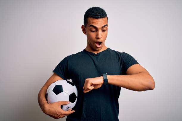 homem afro-americano bonito jogando footbal segurando bola de futebol sobre fundo branco olhando para o tempo de relógio preocupado, com medo de chegar atrasado - football human hand holding american football - fotografias e filmes do acervo