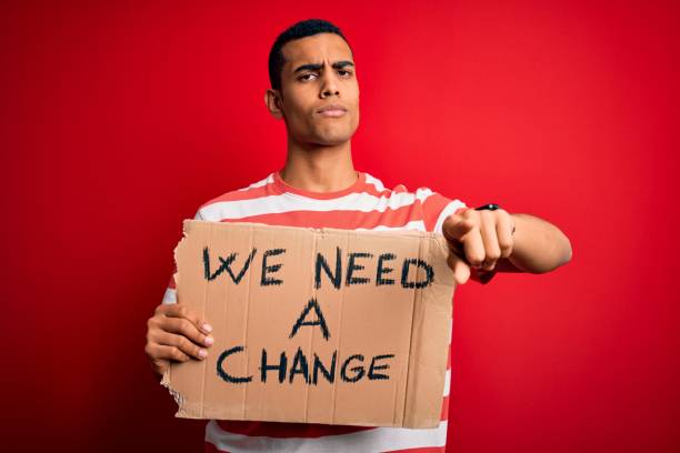 istock Young handsome african american activist man asking for change holding banner with message pointing with finger to the camera and to you, hand sign, positive and confident gesture from the front 1214209843