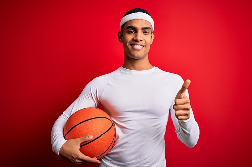 Young handsome african american sportsman holding basketball ball over red background happy with big smile doing ok sign, thumb up with fingers, excellent sign