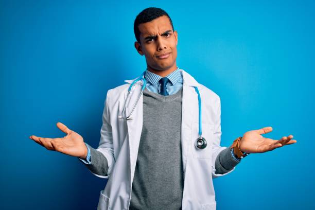 stilig afroamerikansk läkare man bär kappa och stetoskop över blå bakgrund clueless och förväxlas med öppna armar, ingen aning koncept. - doctors talking confused bildbanksfoton och bilder