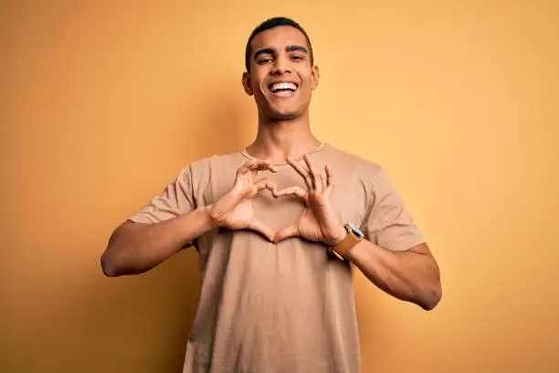 Photo of Young handsome african american man wearing casual t-shirt standing over yellow background smiling in love doing heart symbol shape with hands. Romantic concept.