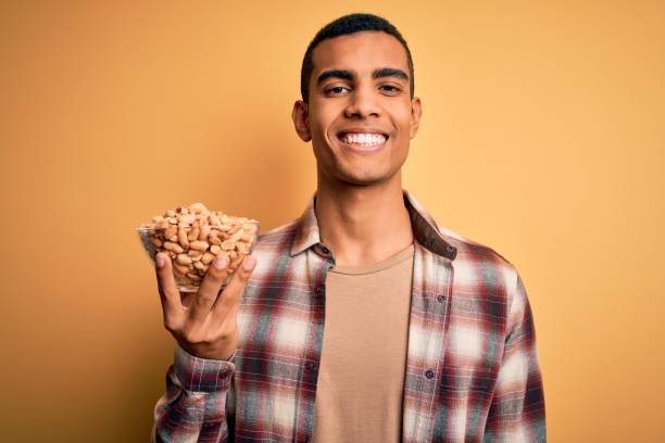 잘 생긴 아프리카 계 미국인 남자 들고 그릇 과 히스 땅콩 위에 노란 배경 에 행복한 얼굴 서서 치아를 보여주는 자신감 미소와 미소 - peanut nut snack isolated 뉴스 사진 이미지