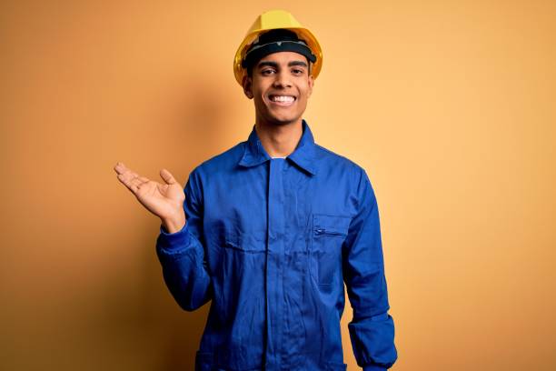 istock Young handsome african american worker man wearing blue uniform and security helmet smiling cheerful presenting and pointing with palm of hand looking at the camera. 1214211803