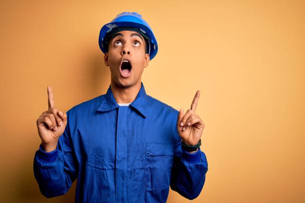 istock Young handsome african american worker man wearing blue uniform and security helmet amazed and surprised looking up and pointing with fingers and raised arms. 1214211945