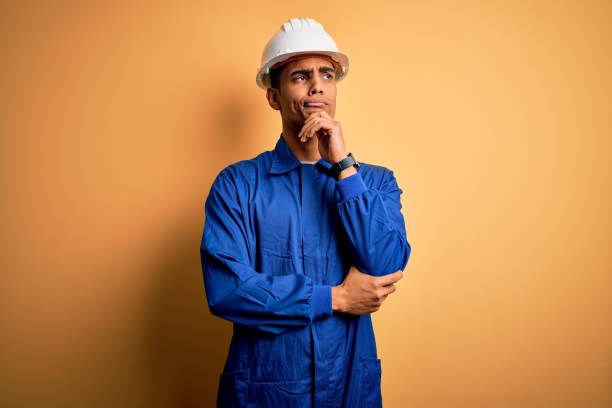 istock Young handsome african american worker man wearing blue uniform and security helmet with hand on chin thinking about question, pensive expression. Smiling with thoughtful face. Doubt concept. 1214212026